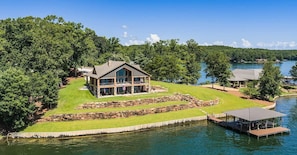 A large, two-story house with a stone foundation and a wrap-around porch, situated on a grassy area with a dock extending into a lake.