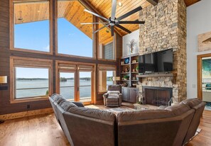A spacious living room with a large fireplace, a ceiling fan, and a view of a body of water through large windows.