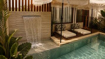 A pool area with a waterfall feature, seating with cushions, and a wooden deck.