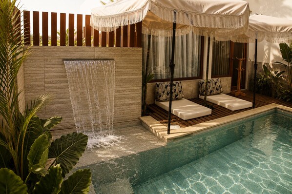 A pool area with a waterfall feature, seating with cushions, and a wooden deck.