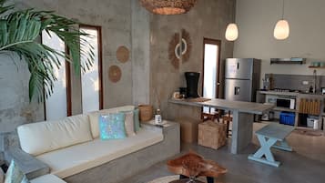 Sala de estar moderna com sofá, mesa de centro e área de cozinha com geladeira e micro-ondas.