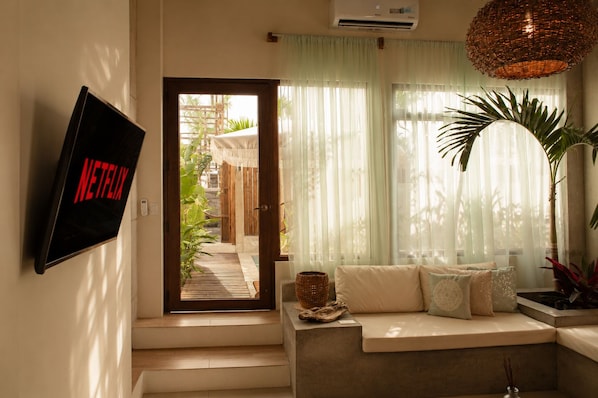 A living room with a sofa, a wall-mounted TV displaying the Netflix logo, a large window with sheer curtains, and a hanging wicker light fixture. - Areia Bali: A/C, private pool, just steps from the ocean (El Paredón Buena Vista)