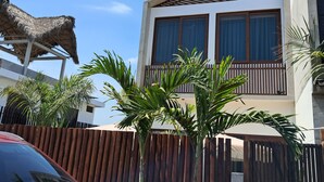 Un edificio moderno de dos pisos con un balcón, rodeado por una cerca de madera y plantas tropicales.