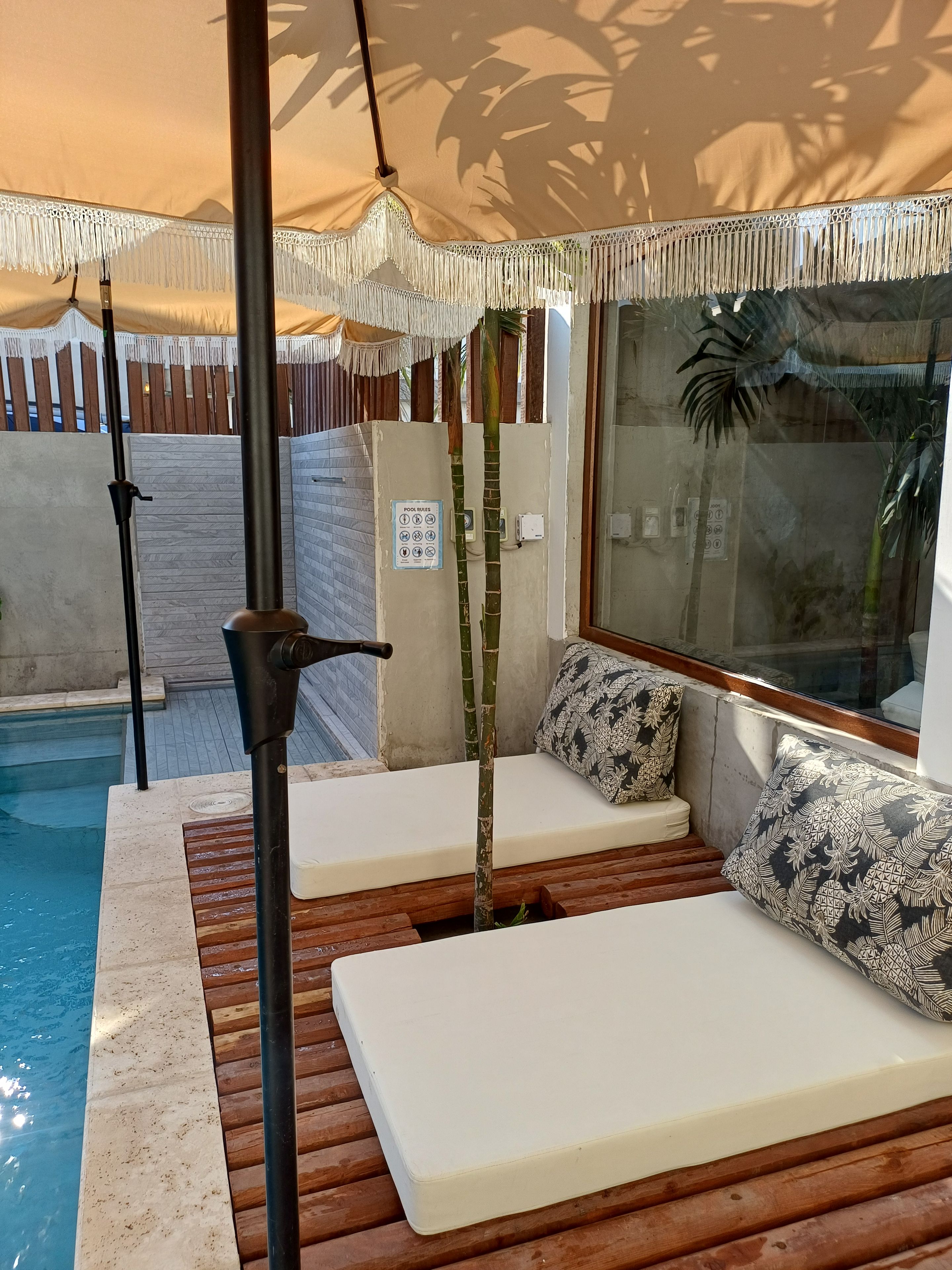 Zona junto a la piscina con terraza de madera, dos sillas reclinables blancas, un pilar de bambú y una piscina con agua cristalina.