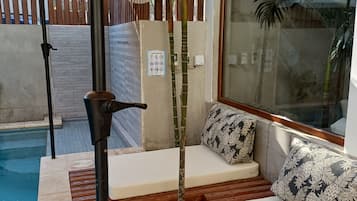 Uma área à beira da piscina com deck de madeira, duas cadeiras de sol brancas, um pilar de bambu e piscina com água cristalina.