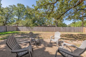 An outdoor seating area with Adirondack chairs and a table, surrounded by a wooden fence and trees. - Montana: Custom Home, 4 BR, Close to Texas A&M (College Station)