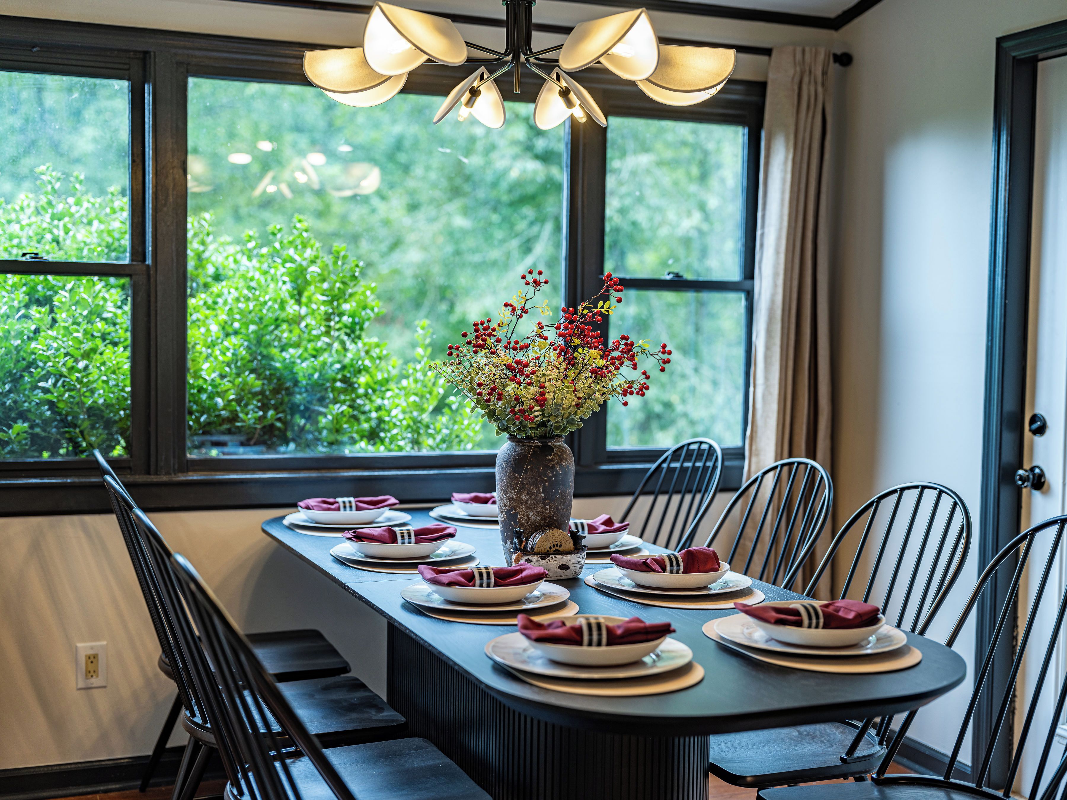 A dining table set with plates, napkins, and a vase with flowers, positioned near a window with a view of greenery.