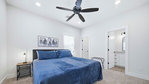 Une chambre à coucher comprenant un lit, un ventilateur de plafond, une table de chevet, une lampe, un tableau, et une salle de bain avec un miroir et des placards.