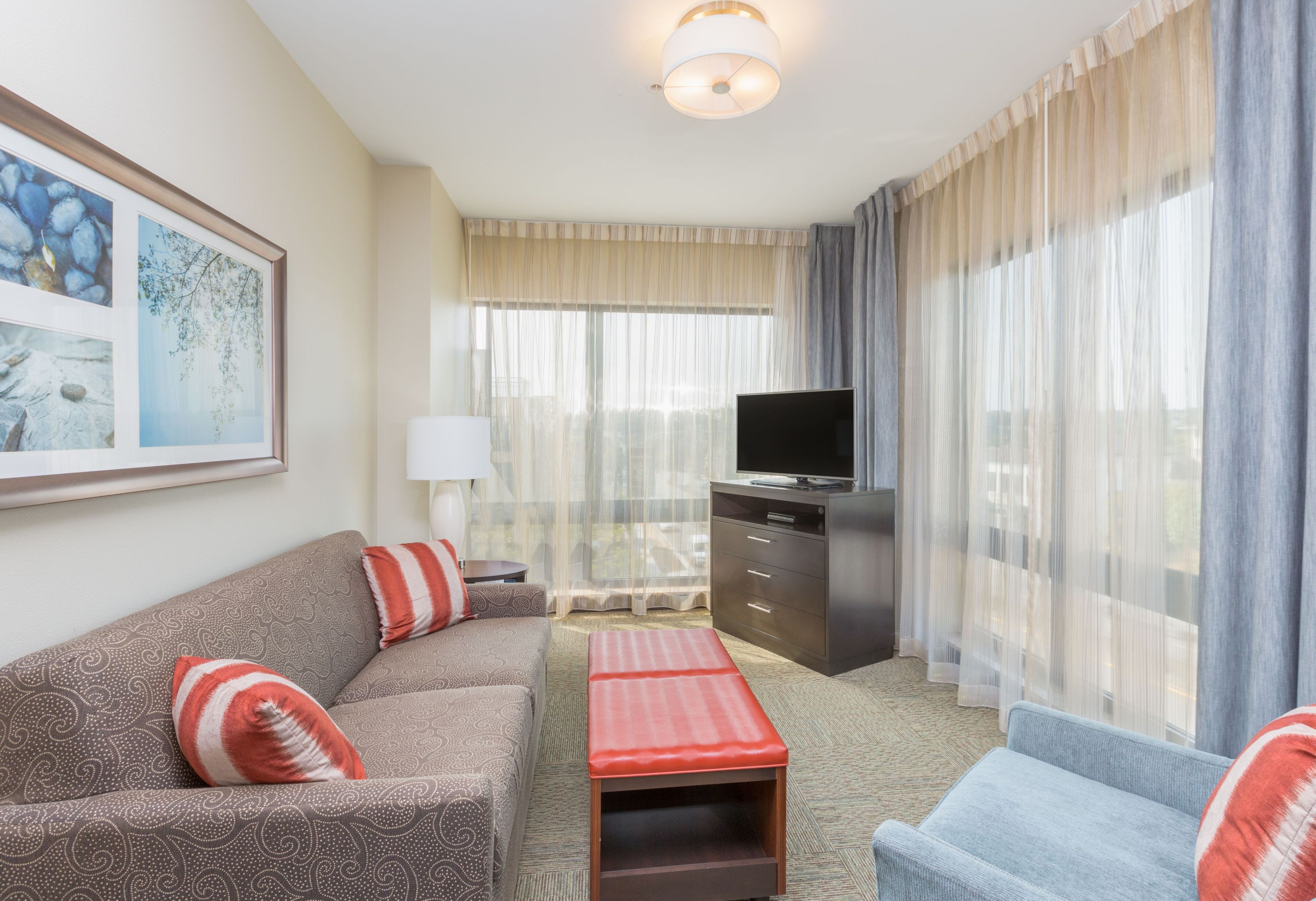 Habitación de hotel con sofá, un puf rojo, un televisor sobre un mueble y un ventanal con cortinas translúcidas.