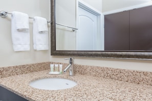 Une salle de bain avec un comptoir en granit, un lavabo blanc, un miroir et trois serviettes blanches suspendues à un porte-serviettes.