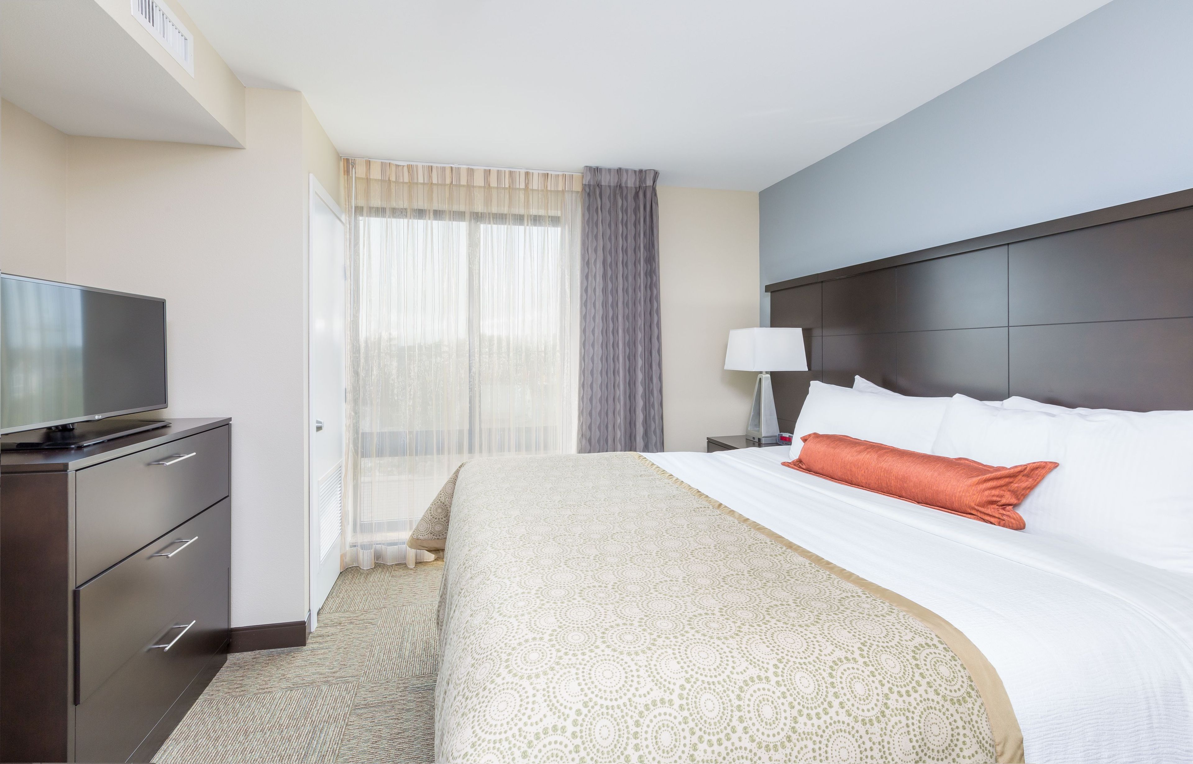 A hotel room with a bed, a TV on a dresser, a lamp, and a window with curtains.