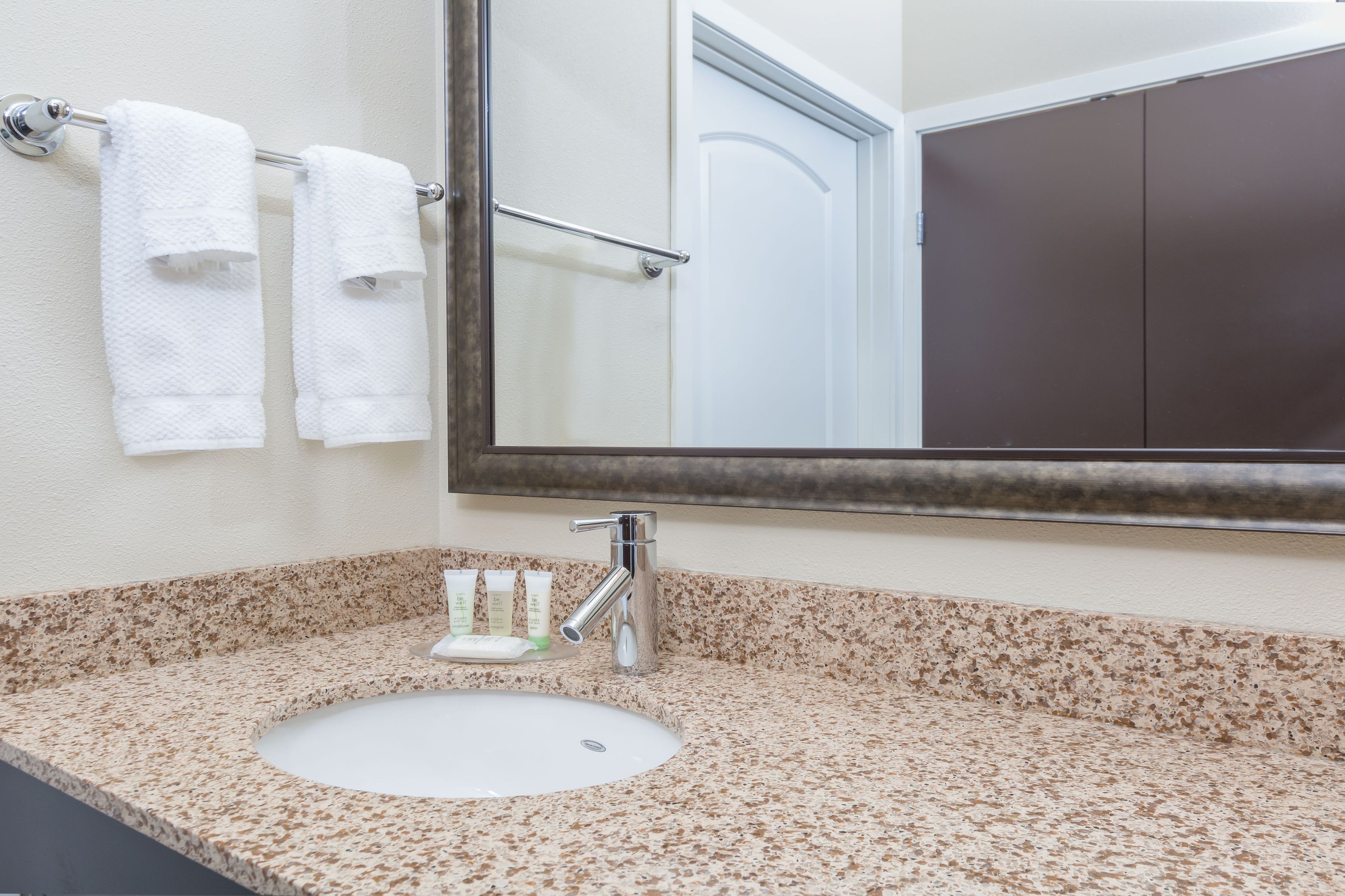 A bathroom with a granite countertop, a white sink, a mirror, and three white towels hanging on a towel rack.