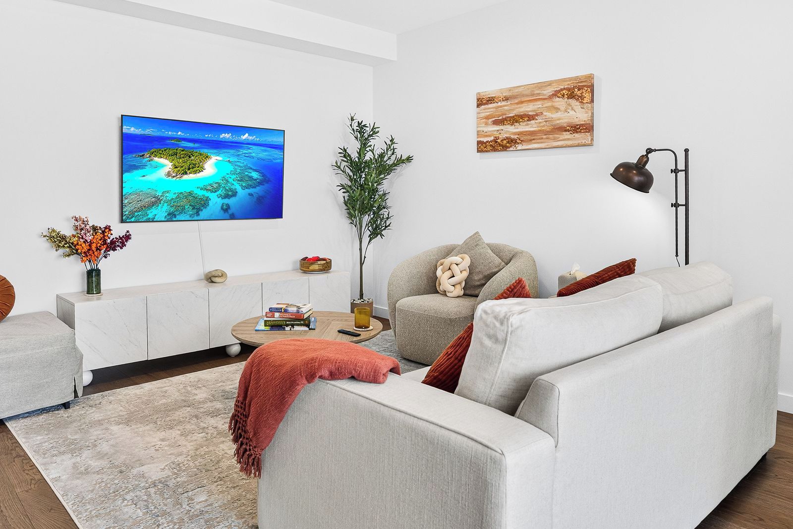 A modern living room with a flat-screen TV, a coffee table with books, a potted plant, and a sofa with cushions.