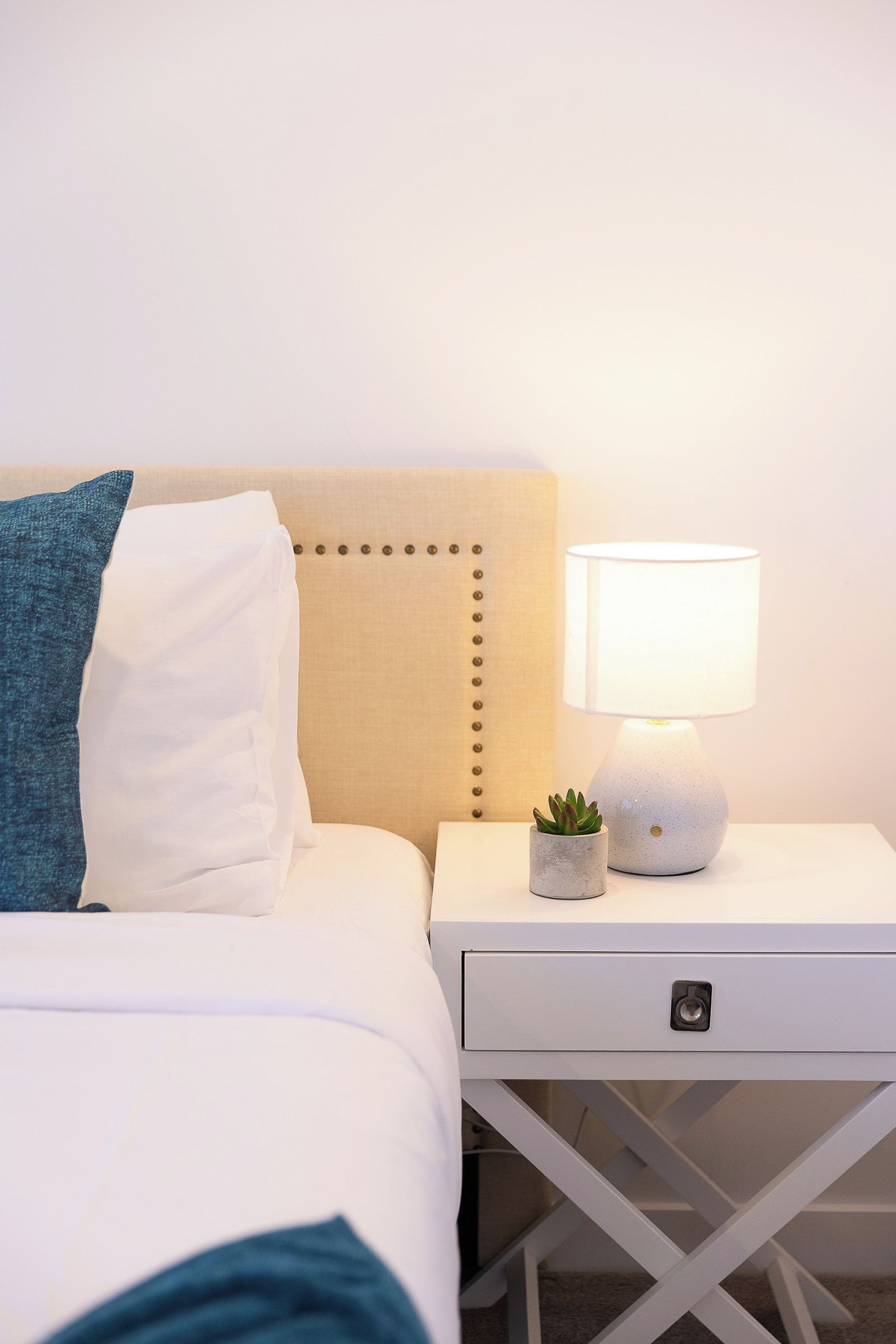 A neatly made bed with a headboard, a bedside lamp, a small potted plant, and a white nightstand with a drawer.