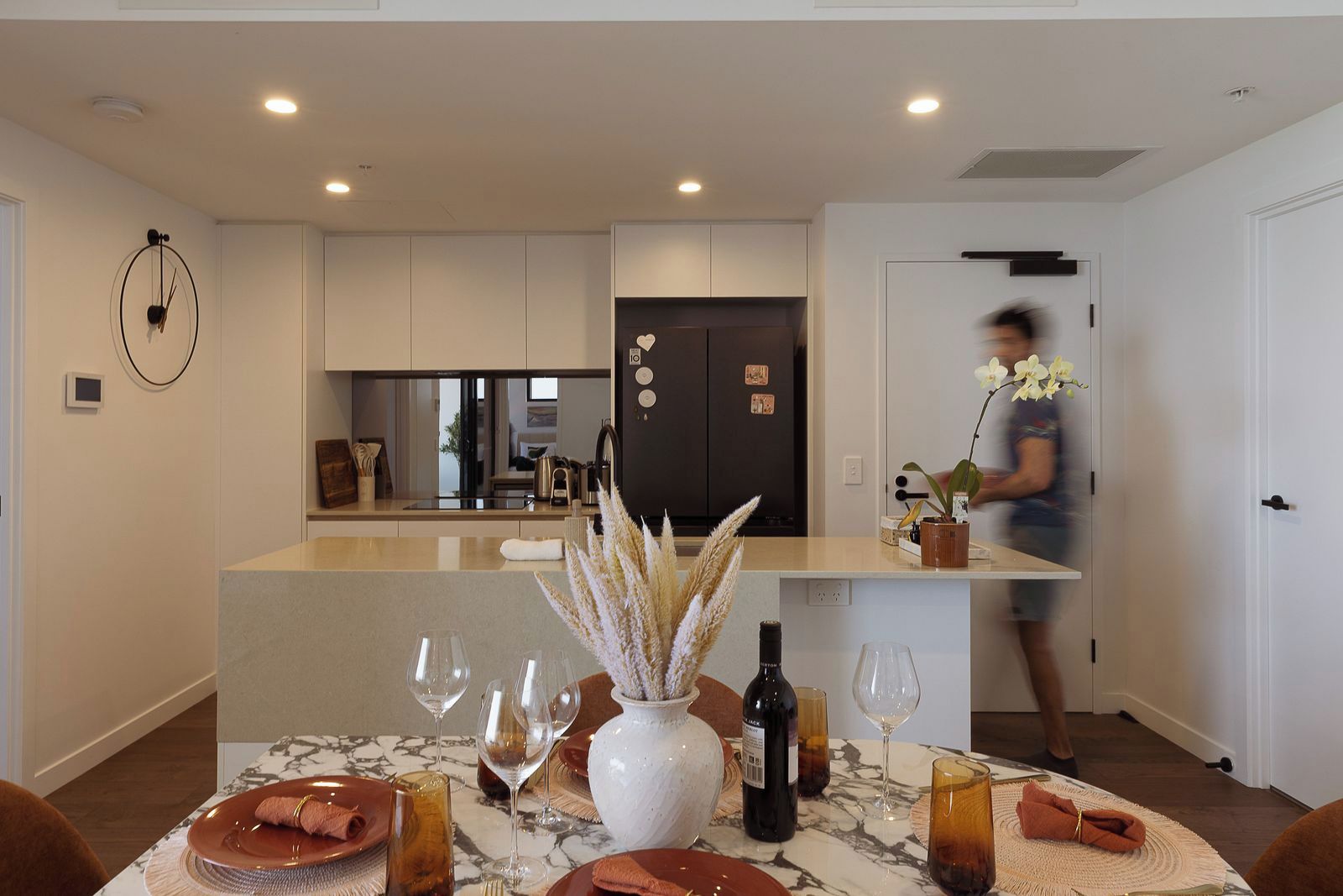 A modern kitchen with a central island, a dining table set with plates, glasses, and a bottle, and a person in the background.