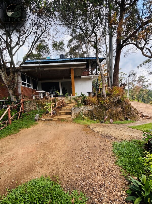 A building with a covered porch, a dirt path leading to it, and surrounding greenery.