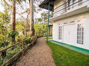 A pathway leading to a white building with a balcony, surrounded by greenery and trees.