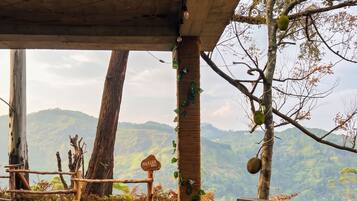 A rustic outdoor seating area with a wooden table and chairs, surrounded by plants and a view of a hilly landscape.