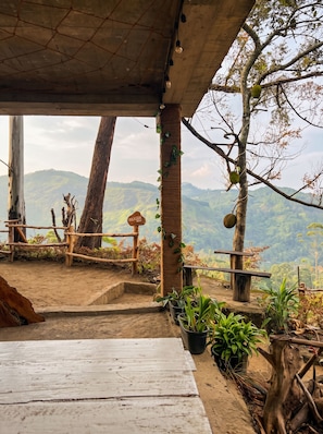 A rustic outdoor seating area with a wooden table and chairs, surrounded by plants and a view of a hilly landscape.