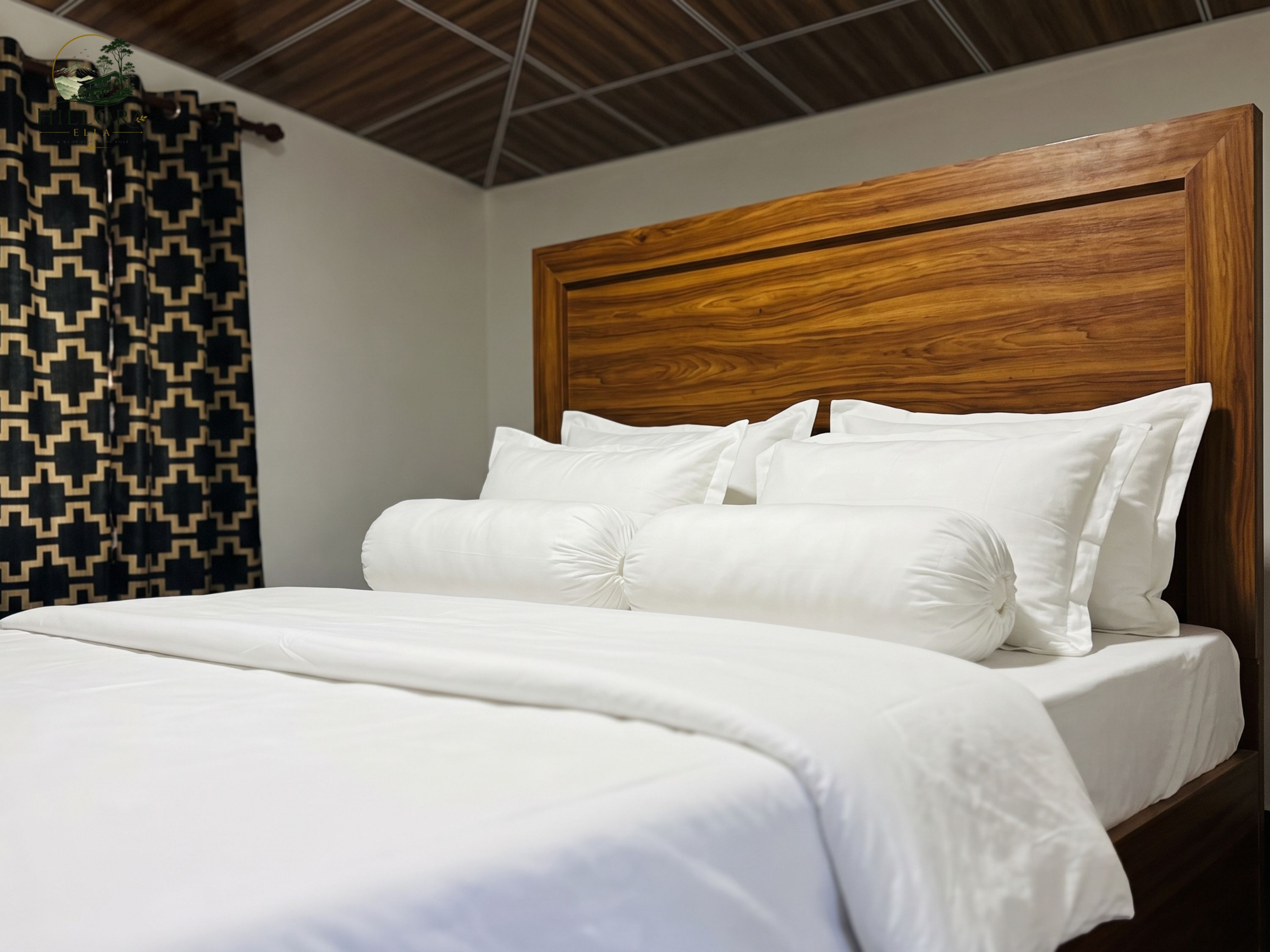 Una cama con ropa blanca y un cabecero de madera en una habitación con cortina estampada.