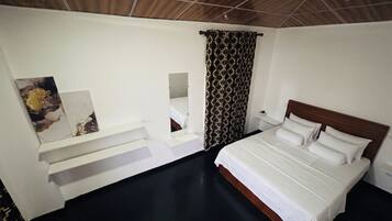 A hotel room with a bed, a mirror, a shelf with artwork, a patterned curtain, and a wooden ceiling.