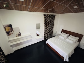 A hotel room with a bed, a mirror, a shelf with artwork, a patterned curtain, and a wooden ceiling.
