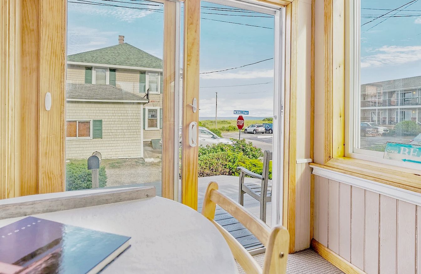 A room with a table and chairs, a window with a view of a street and houses, and a stop sign in the distance.