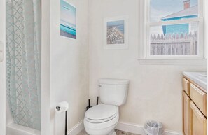 A bathroom with a toilet, a shower curtain, and a window with a view of a beach and a fence. - The Sea Gaze - 4BR with Beach Access (Old Orchard Beach)