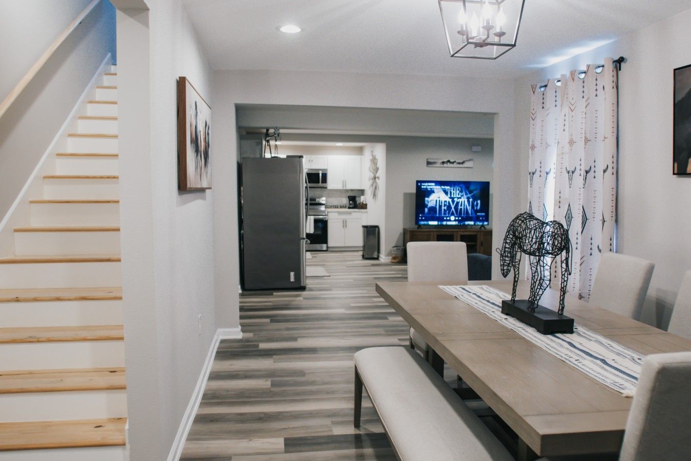 A modern living space with a staircase, a dining area with a bench, a kitchen with a refrigerator, and a television displaying 'The Expan'.