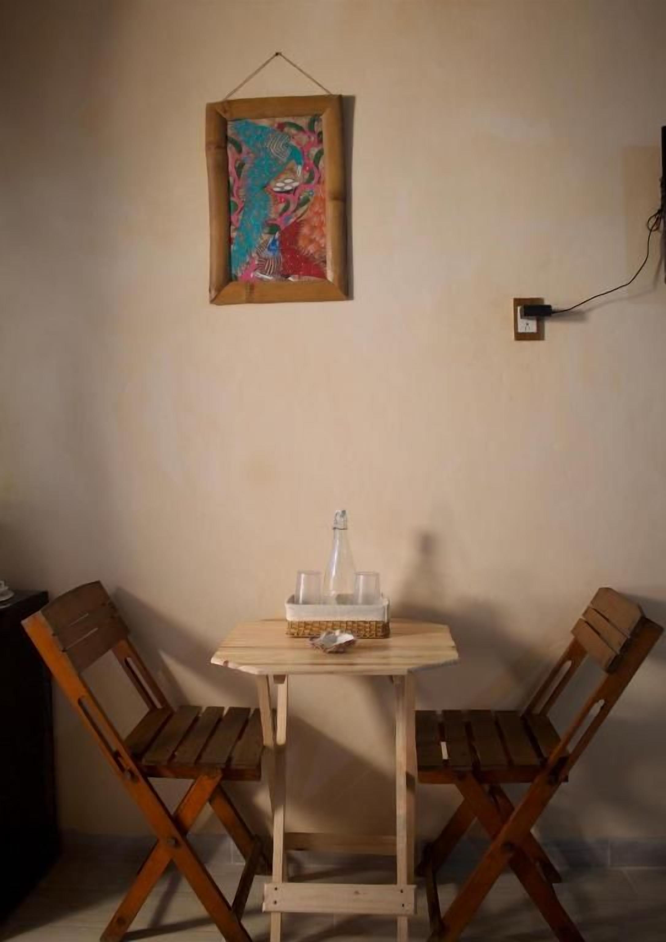 A small wooden table with a bottle and glasses, two wooden chairs, and a framed artwork on the wall.