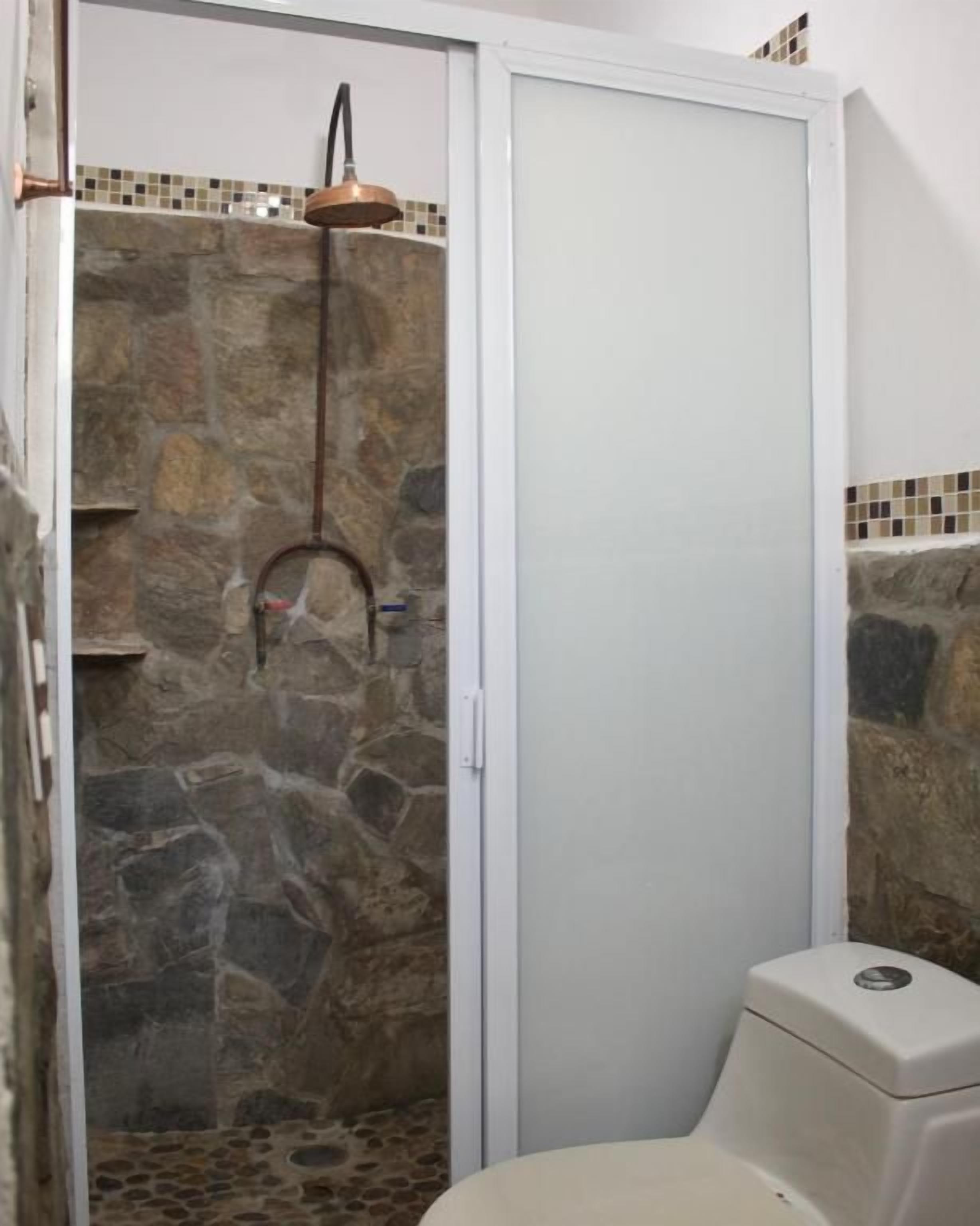 Une salle de bain avec une douche en pierre, des toilettes blanches et un mur recouvert de carreaux derrière le lavabo.