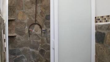 Une salle de bain avec une douche en pierre, des toilettes blanches et un mur recouvert de carreaux derrière le lavabo.