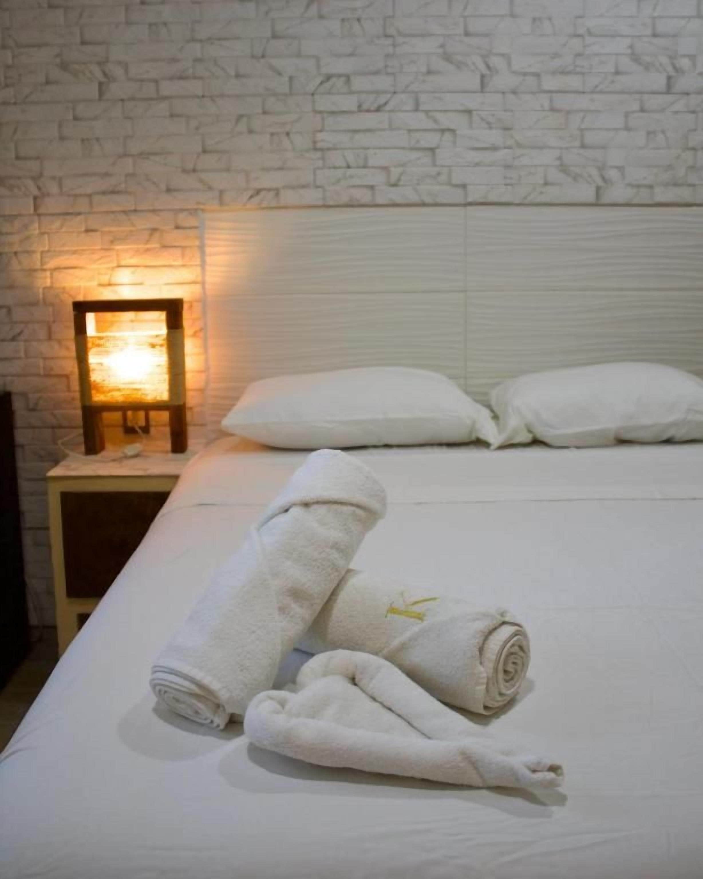 Una habitación de hotel con pared de ladrillo blanco, una cama con ropa de cama blanca, dos almohadas, una mesita de noche con una lámpara y dos toallas enrolladas.