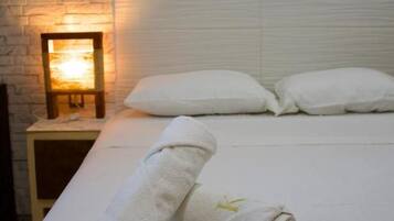 Una habitación de hotel con pared de ladrillo blanco, una cama con ropa de cama blanca, dos almohadas, una mesita de noche con una lámpara y dos toallas enrolladas.