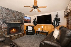 Un salon chaleureux avec une cheminée en pierre, des meubles en cuir, un ventilateur de plafond et un téléviseur fixé au mur.