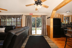 Una habitación con sofá de cuero negro, ventilador de techo de madera, alfombra negra, área de cocina con armarios de madera y vista por una puerta de vidrio.