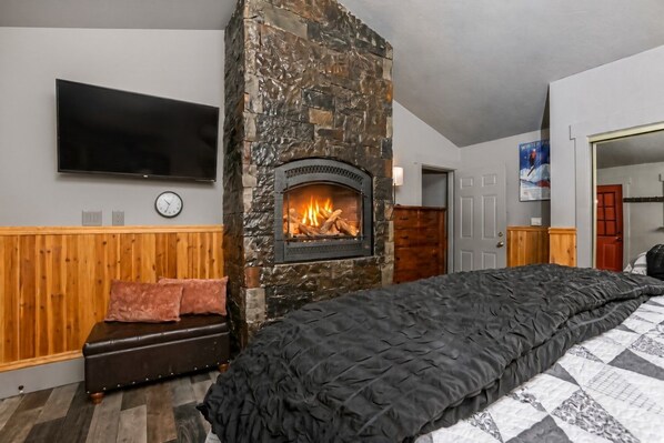 A bedroom with a stone fireplace, a bed with a black quilt, a wooden bench, a television mounted on the wall, and a clock.
