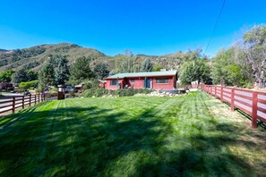 A red building with a green roof, surrounded by a wooden fence and a well-maintained lawn. - Cozy, Family Cabin-Style Home, Walk to River & Downtown, Dog Friendly (Glenwood Springs)