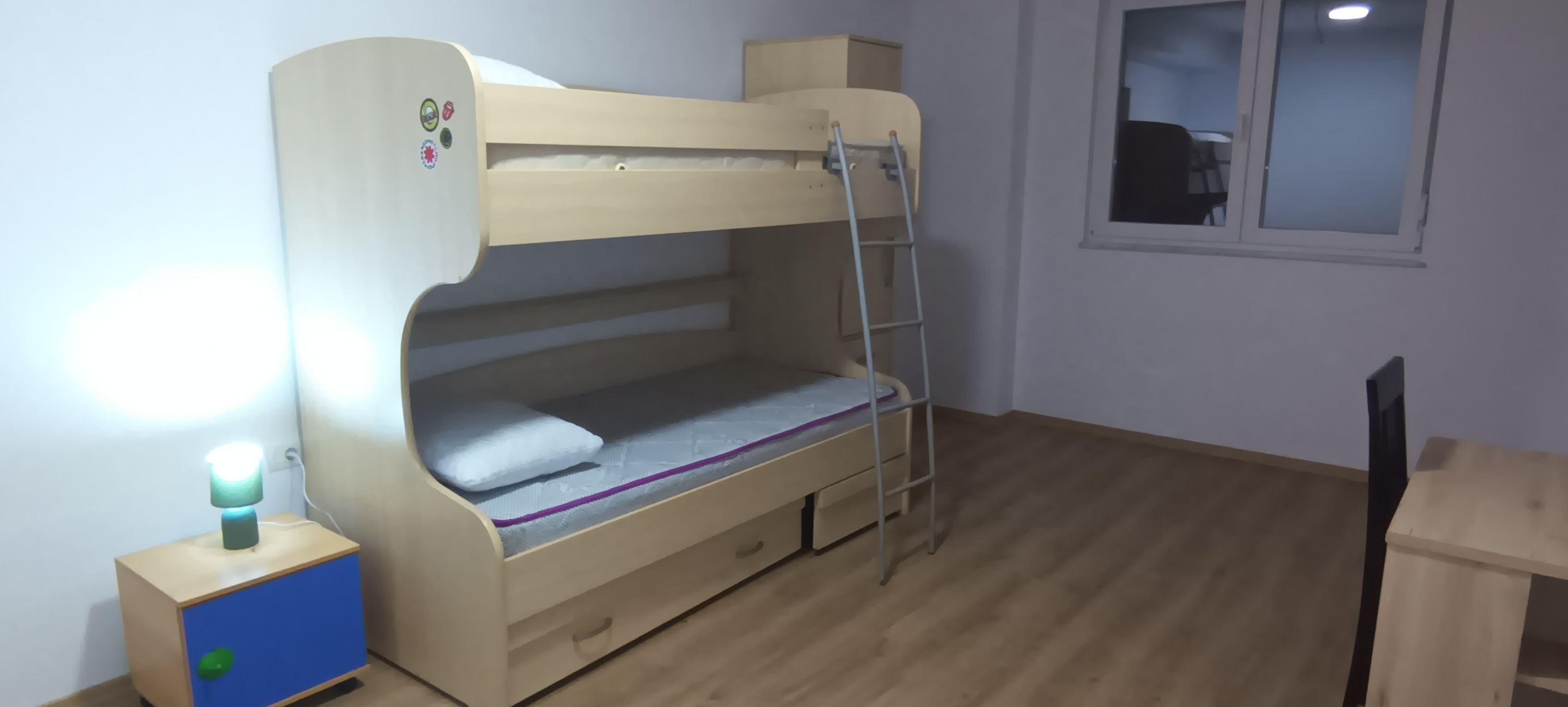 A bunk bed with a ladder, a desk, and a window in a room with wooden flooring.
