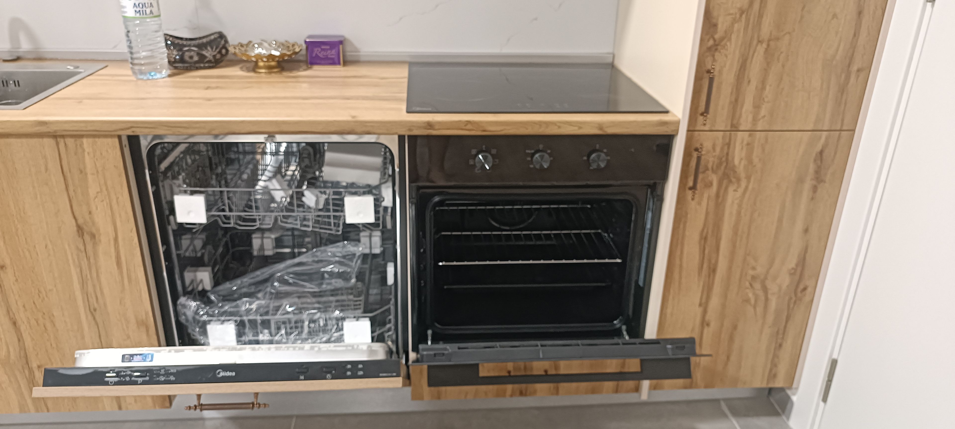 Una cocina con lavavajillas y horno integrados, armarios de madera y una encimera con una botella de agua y objetos decorativos.