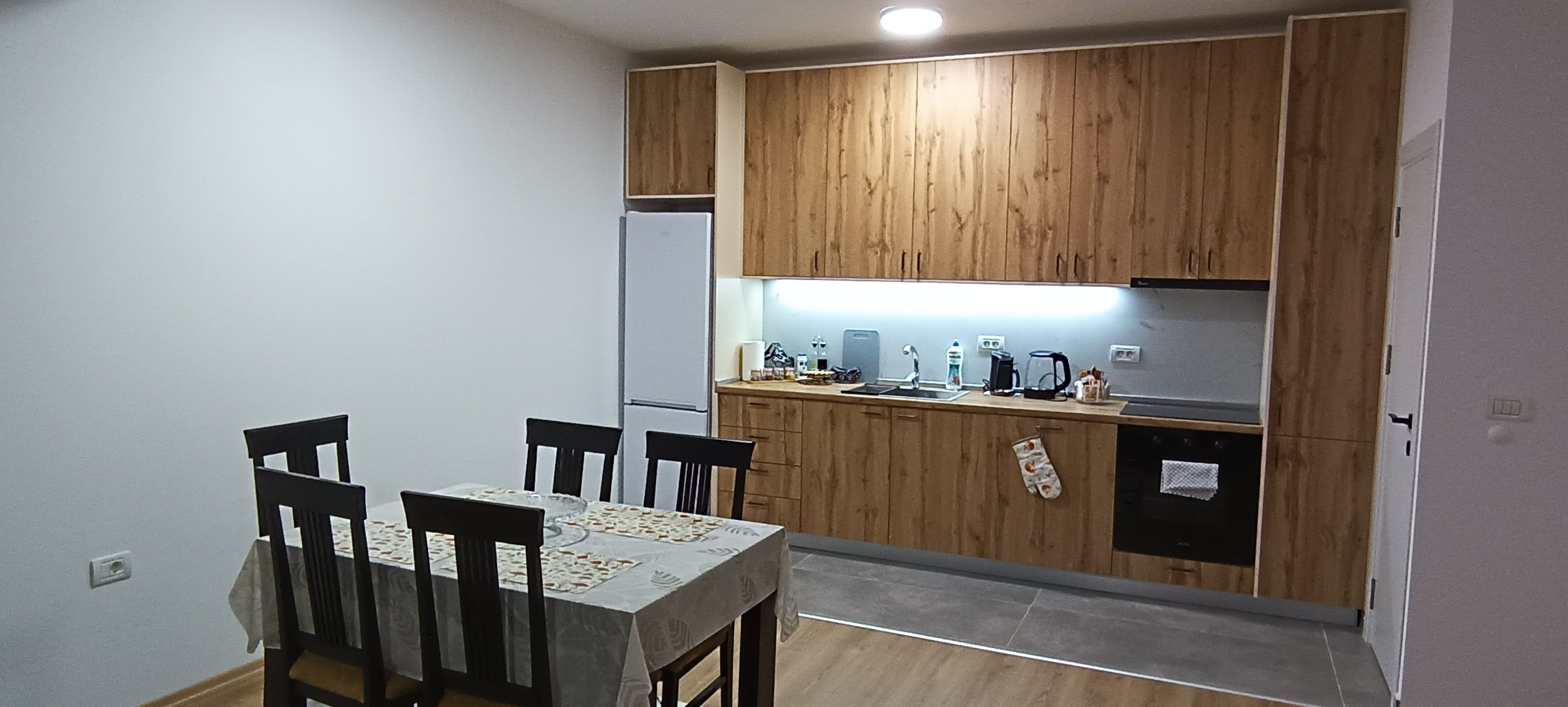 Een compacte keuken met houten kastjes, een eettafel met stoelen en een aanrecht met diverse apparaten.