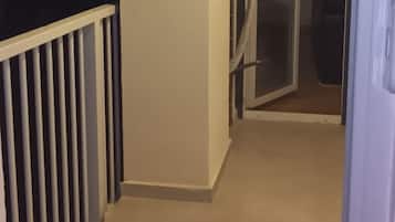 A narrow corridor with a white railing on one side and a door at the end leading to a room with a bed and a nightstand.