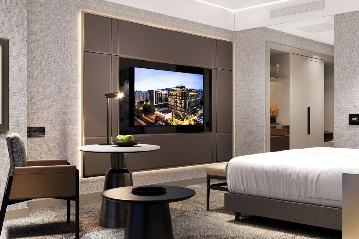 A modern hotel room with a large bed, a TV mounted on the wall, a small round table with a bowl of fruit, and a chair.