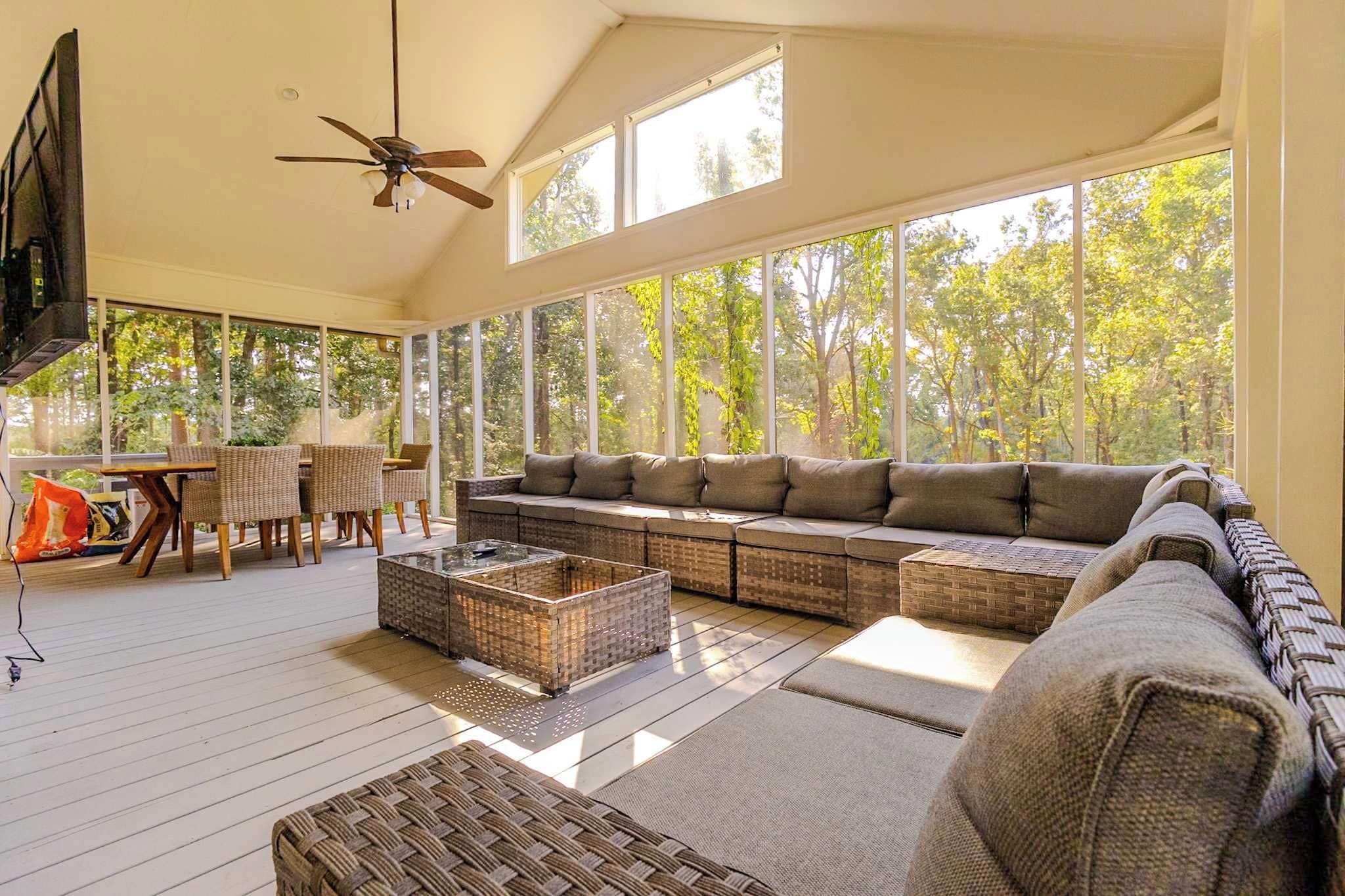 A spacious living room with a large sofa, a coffee table, and a TV. There are windows providing a view of trees outside.