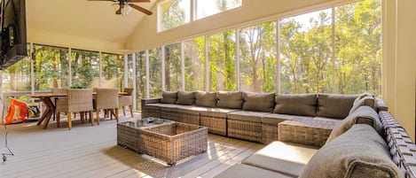 A spacious living room with a large sofa, a coffee table, and a TV. There are windows providing a view of trees outside.