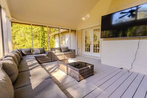Ein sonnendurchfluteter Außenbereich mit einer Couch und Sesseln, einem Couchtisch aus Glas und einem an der Wand befestigten Fernseher.