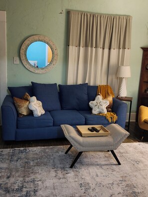Ein Zimmer mit einem blauen Sofa, einem runden Spiegel, einem Holznachttisch und einer Lampe.