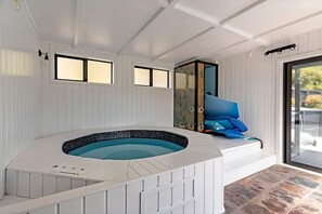 A hot tub with a stone-like floor, white walls, and a glass door leading outside. - Peaceful Upper Hutt Hideaway • Pool • Spa • Sauna (Upper Hutt)