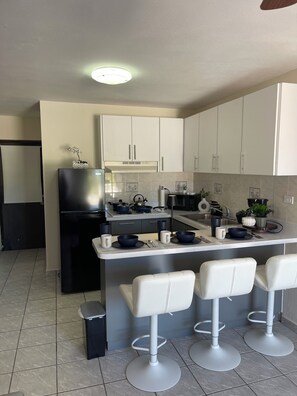 A modern kitchen with white cabinets, a black refrigerator, a central island with bar stools, and a ceiling light. - Cozy 2BR in Carolina 12min to Beach & Airport Near All (Carolina)