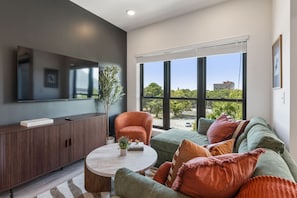 Ein modernes Wohnzimmer mit einer Couch, einem Sessel und einem Couchtisch. Es gibt ein großes Fenster mit Jalousien, einen am Wand montierten Flachbildfernseher und einen Blumentopf.